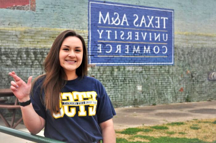 Picture of Heather Rodriguez making the lion sign with behind a mural of 皇冠体育365赌博&M University Commerce.